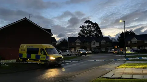 A cul-de-sac at dawn, cordoned off with blue and white police tape. To the left is a street sign for Allerton close, covered in police tape. A large yellow police van sits behind the cordon to the left, behind is a smaller police van in the center of the street, to the right is a police car.