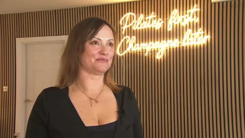 A woman stood in front of a wall with a sign saying 'Pilates first, champagne later', looking next to the camera