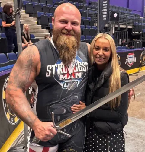Supplied A man in a dark leotard, with a bald head and long brown beard is holding a sword. His leotard has the words "UK Strongest Man Live" printed on it. He is hugging a woman with long blonde hair and a black jacket.