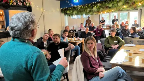 A large group of people are seen in a room sitting at tables as they listen to a woman who is standing up and speaking. The speaker is facing away from camera and has a green jumper