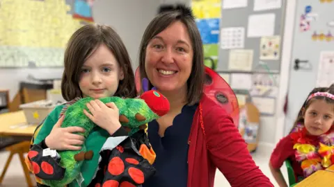 Murphy, a P2 schoolboy, poses for a photo with his teacher Mrs McIntyre, a woman with shoulder-length dark hair.  Murphy has dark hair and is wearing a green t-shirt under a set of colourful, spotted fabric wings. He is hugging a stuffed toy caterpillar.  His teacher is wearing a red cardigan and red and black ladybird wings.   They are sitting in a bright classroom.