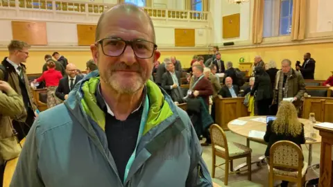 A man standing in a green jacket. Behind him, a council meeting has just taken place. 