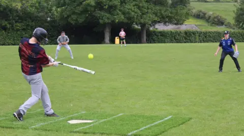 GB Softball A few boys are playing softball on a green pitch with trees in the background. They are wearing long trousers and T-Shirts.