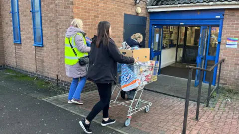 Liam Barnesand One person pushing a trolley full of bags and boxes of food. Two other people can be seen behind. They are walking towards a blue doorway.
