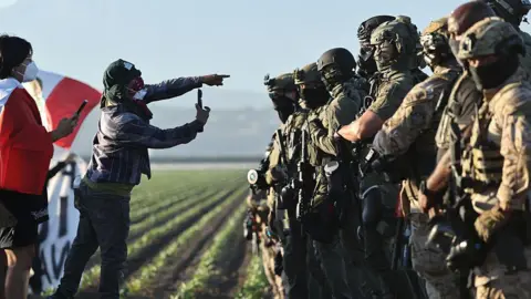 Two protesters in dust masks film federal troops in gas masks in a field of crops in Southern California. One protester flies a Mexican flag
