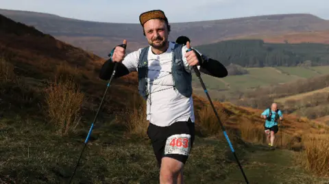 Shelly Price A man in a white top running up a steep hill, he is holding two poles and wearing a cap. 