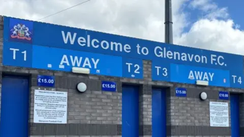 A picture taken outside Glenavon Football Club in Lurgan. There is a 'Welcome to Glenavon FC' sign, with four ticket booths within the grey brick wall.