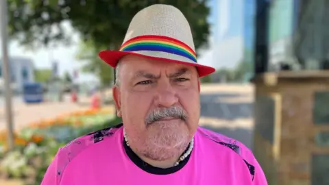 A man, wearing a trilby, on his head, with a striped band on it. He is wearing a pink T-shirt. He is in front of a street, with vehicles in the distance, a brick wall and trees, that are all slightly blurred.
