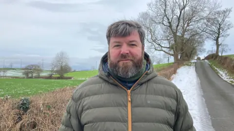 Jamie Corry looking into the camera with fields and a road behind him. The fields and road have a small amount of snow on them. 