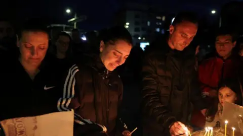 Four adults and a child stands in lighting candles. Others can be seen behind. The picture has been taken at night and is dark