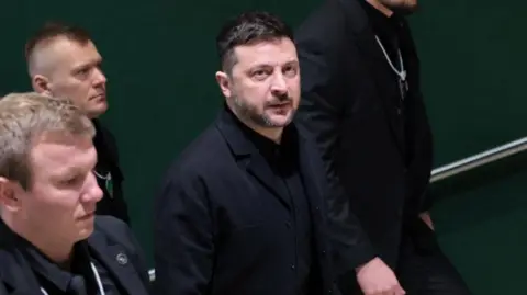 A man wearing a black jacket and shirt surrounded by three other men in black rides up an escalator
