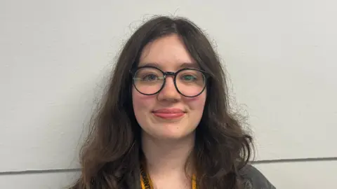 George King/BBC A head and shoulders image of a young woman wearing glasses and standing in front of a white wall. She is smiling and looking into the camera. 