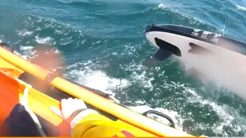 RNLI A blue and white paddleboard in the sea next to a lifeboat, with a crew member's hand visible