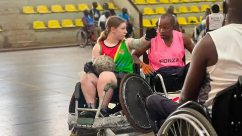 Sarah Burrows (left) in conversation with a man in a sports hall. They are both in wheelchairs. Sarah has a round ball in her lap. She has brown hair, tied back, and is wearing a red and green top. The man next to her has short black hair and is wearing a pink top.