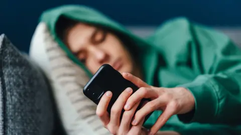 Getty Images A teenager wearing a green hoody with the hood pulled up, lies on cushions on a sofa looking at his mobile phone