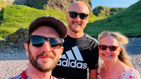 Kieran Hutcheon, Dan Bickmore and Karen Gill, they are smiling at the camera at a beach setting.