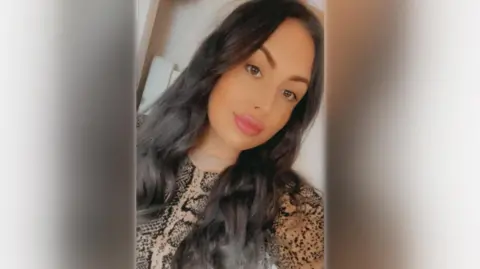 Family handout A young woman with long dark brown hair and brown eyes in a selfie picture. She is looking off to the side of the frame and is wearing a snakeskin patterned jumper and pink lipstick. 