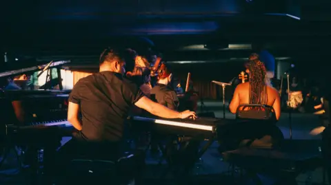 Kieran Irvine Musicians are facing away from the camera in a dark room. There is a man playing the keyboard in the foreground of the image