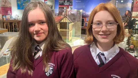 Two girls wearing school uniform of maroon v-neck sweater over a white shirt and striped black, red and silver tie. Aoife on the left has long brown hair and Olivia on the right has an auburn bob and round glasses.