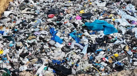 A huge pile of rubbish dumped in a three-sided pit. Two people can be seen in the background, along with several vehicles and containers.