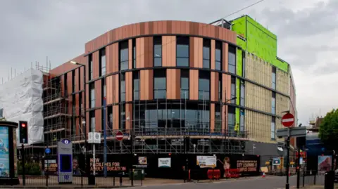 The large orange and black curved building stands on a corner. It has a green and gold section on the right hand side of the image. There are some advertising hoardings and small scaffolding at ground level.