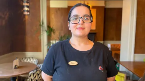 BBC/Mark Ansell A woman with dark hair, pulled back and black rimmed glasses, smiles at the camera. Her arms are neatly placed behind her back and she is wearing a black top