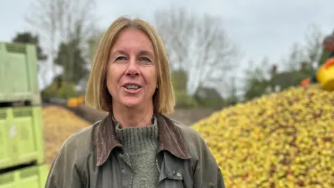 A woman in a green jacket and jumper stands in front of a large pile of yellow apples and green containers. She has blonde hair past her ears, a slight open-mouthed smile on her face and wears a light green jumper under a green wax-style jacket.