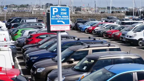 There is a harbour in the background, with masts of yachts just behind a sea wall. in the foreground there is a full car park with differently coloured cars, including red and green ones.
There is a car parking sign in the foreground which tells motorists the park is a 10 hour disc zone.