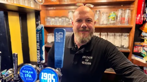 Jonathan Kaye standing at the bar of the Prince of Wales pub, wearing a branded black top.