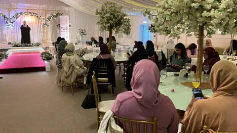 Nicola Haseler/BBC A view of the stage from the back of the room. A woman stands at the podium addressing the crowd, who sit at tables around the room. Some are looking at their mobile phones. The majority of the women wear the hijab.