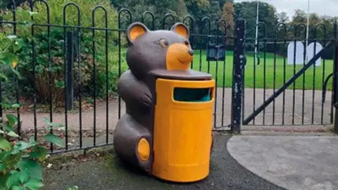 Andy Bailey A yellow bin, which is shaped as a bear holding a barrel. There is a fence with a gate in the background. The bin is sat on a tarmac path.