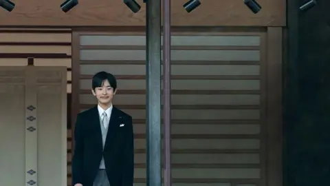 Getty Images Prince Hisahito smiles and stands behind a window. He is wearing a black suit.
