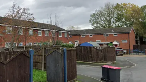 The image shows the scene where a man was attacked. It is a cul-de-sac of relatively modern red brick houses, with fences of about six-feet obscuring views of the gardens. Behind one fence can be seen what looks like a blue police forensic tent. There are two black, pink-lidded wheelie bins on the road in the foreground. 
