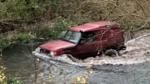 A red 4x4 drives along a section of a Hampshire chalk stream open to traffic