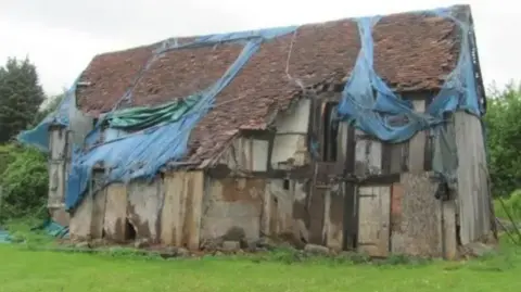 One of the dilapidated buildings at St Michael’s Place, Saltisford, Warwick. It is made from stone and timber and has a blue covering that has worn away in many places. It has grass in front with a tree behind the building.
