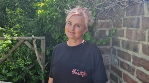 Supplied Sue Davison, who has cropped blonde hair which has streaks of pink. She is wearing a black t-shirt with This life Collective written on the left pocket in pink cursive writing. She is standing in a garden with a brick wall and hedges on either side. 