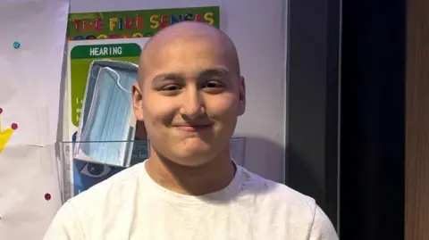Ethan is standing with a toothless grin for the camera. He has on a white t-shirt and has close cropped hair. It is a head shot of him.