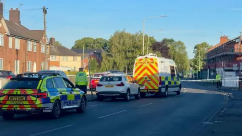 A police car and collision investigation unit van parked outside a police cordon in Gipsy Lane in Leicester. An officer can be seen inside the cordon, which extends across the width of the road, in the background of the image. A second officer wearing a hi-vis jacket is standing in front of the parked police car in the foreground.