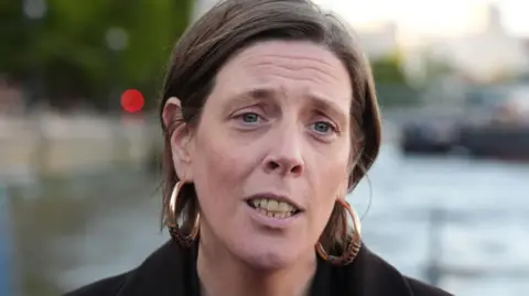 Jess Phillips, who has short brown hair and wears large gold hoop earrings, has a concerned expression as she speaks to the press during a previous press briefing in London