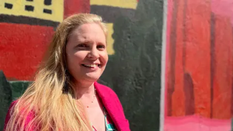 Sue Dougan/BBC Head and shoulders image of Lara Davenport-Ray standing in front of a colourful wall. She has long light hair, is wearing a pink jacket and turquoise dress,