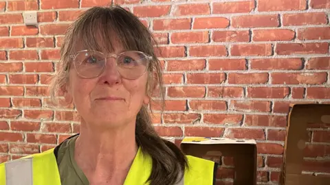 Leigh Milner/BBC Jo Hare, who has long brown hair in a ponytail and is wearing a green T-shirt with a hi-vis vest over the top. She has glasses with a transparent frame and is smiling in front of a brick wall.