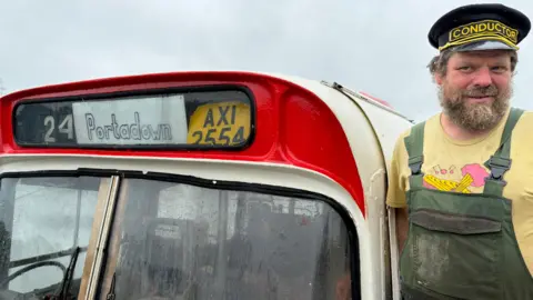 Paddy Bloomer stands on the deck of his bus boat. He is wearing green dungarees over a yellow T-shirt and a black peaked cap with "conductor" written on it in gold font. He has a bushy beard. The bus's destination sign reads "24 Portadown" and beside it is a licence plate reading "AXI 2554".