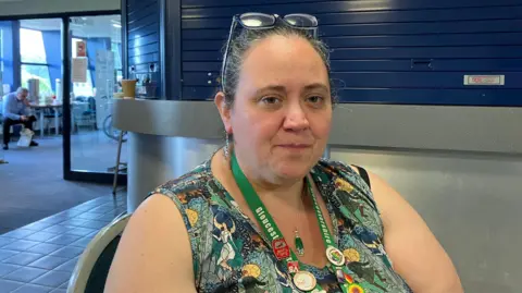 Becky Pain-Tolin sat down with a colourful dress and a green lanyard around her neck. She has glasses on her head. 
