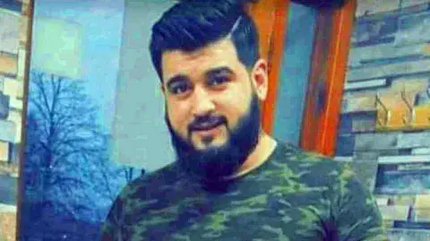 Anas Almarjeh smiles into the camera - he is wearing a T-shirt with military camouflage, while standing indoors. He has dark hair and a beard.