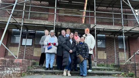 Campaigners stand on the steps at the front of St Joseph's Catholic Church which is made of red brick and covered in scaffolding