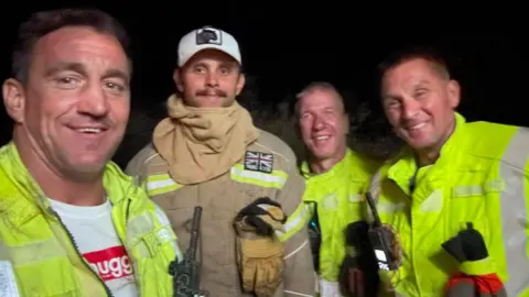Four firefighters from Truro Community Fire Station smile at the camera while working in their high vis uniform at night.