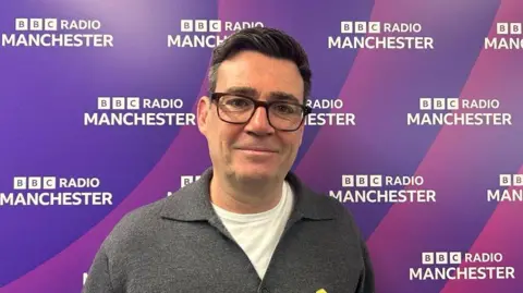 Andy Burnham, 56, who has black hair with streaks of grey and wears black framed galesses, stands in front of a purple screen bearing the BBC Radio Manchster logo. He is wearing a grey button up jumper over a white t-shirt. 