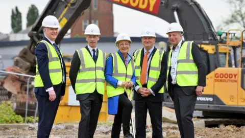 Essex and Suffolk Elective Orthopaedic Centre Four men and one woman wearing high-vis and white hard hats stand on a building site. All are smiling. Earth-moving equipment can be seen behind them.