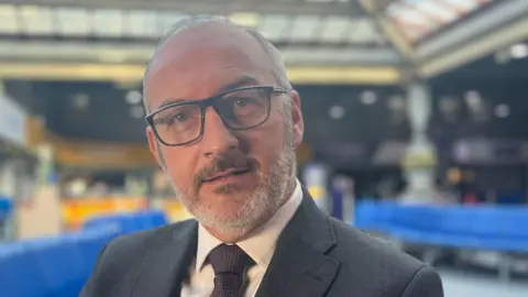 BBC Brittany Ferries and Condor Ferries chief executive Christophe Mathieu sits on a blue bench in Jersey Port. His head is leaning slightly towards his right and he has large square-rimmed glasses. He is wearing a suit and a purple tie.