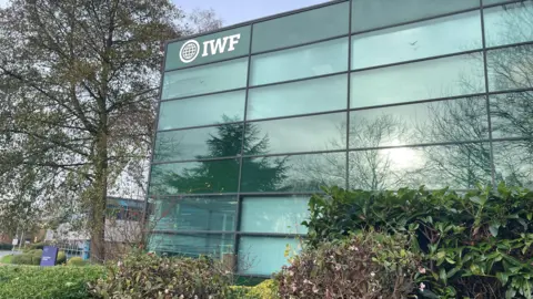 BBC Glass-fronted office block with IWF and a globe sign written in the top-left corner, around the office are other buildings and trees and bushes.